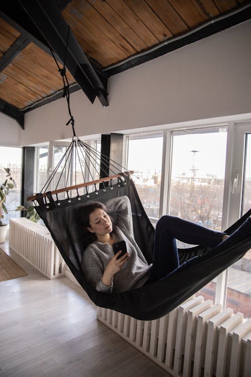 Dreamy female in casual clothes surfing mobile phone while resting in hammock inside of apartment living room