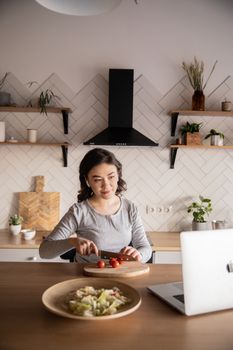 一名妇女在现代厨房里边切蔬菜边用笔记本电脑看食谱