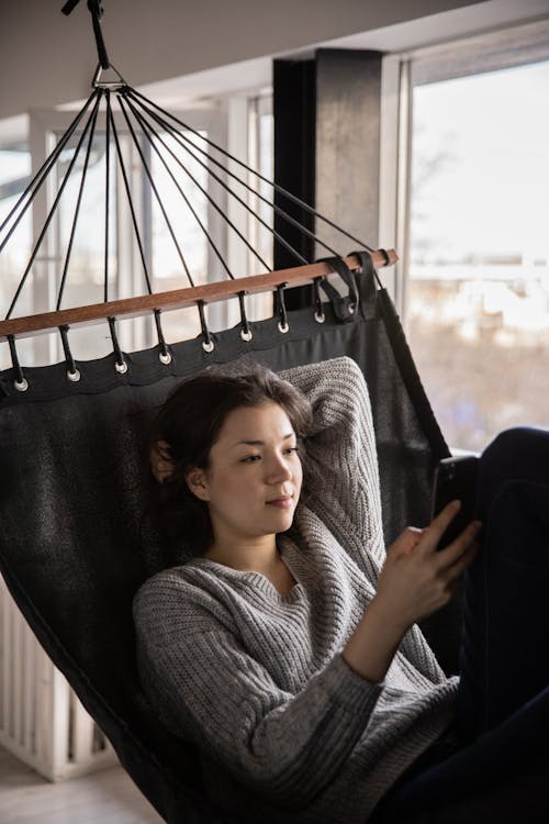 Calm woman chatting on smartphone lying in hammock at home