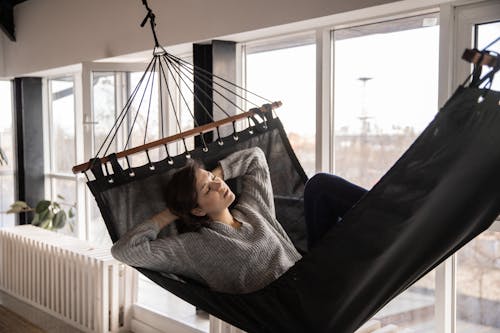 Young lady in casual wear lying in comfortable hammock leaned on hands near window and radiator in apartment in afternoon