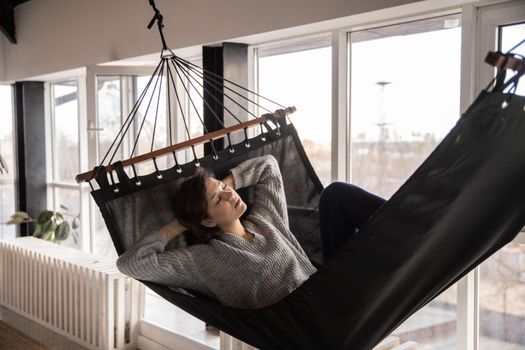 下午，一位穿着休闲装的年轻女士躺在舒适的吊床上，双手靠在公寓窗户和暖气片附近