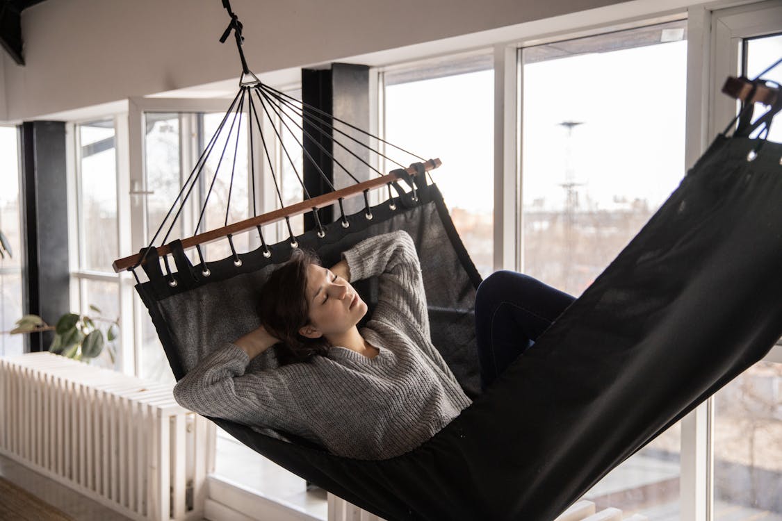 Free Young lady in casual wear lying in comfortable hammock leaned on hands near window and radiator in apartment in afternoon Stock Photo