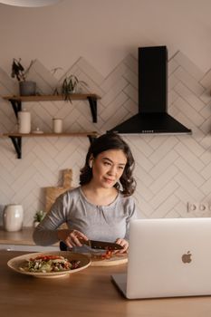 Concentrated young ethnic female cutting fresh vegetables on wooden cutting board while sitting in kitchen in front of open netbook in daylight