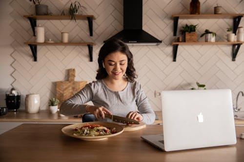 Free Cheerful ethnic female cutting fresh vegetables on cutting board while sitting at wooden table in kitchen with open portable computer in apartment Stock Photo