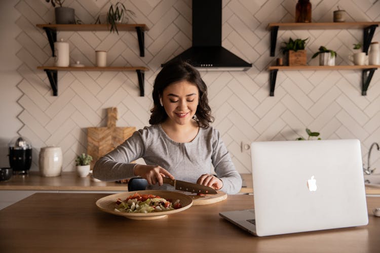 Ethnic Woman Preparing Vegetable Salad In Front Of Open Laptop