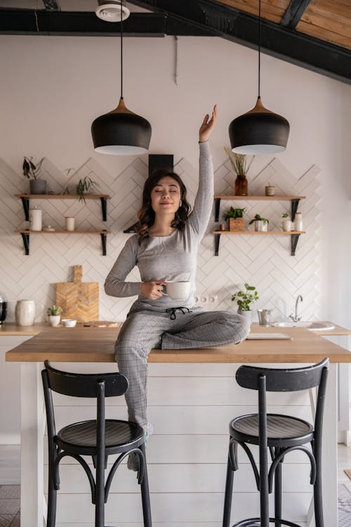 Free Dreamy young ethnic female with closed eyes in pajamas sitting with crossed legs on kitchen table with cup of hot drink and raised hand Stock Photo