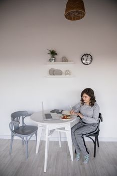 Ethnic female freelancer in casual wear writing in copybook while sitting in front of open laptop at plastic table with salad plate near white wall with clock