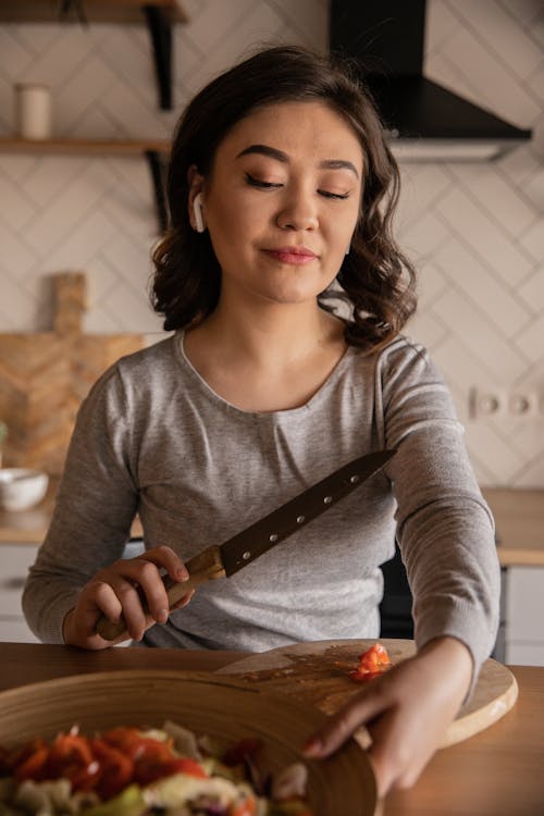 Free Content young ethnic female cook in casual clothes sitting at kitchen table with knife and cutting board while preparing vegetable salad for lunch at home Stock Photo