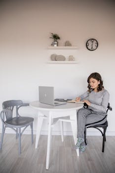 Serious young ethnic female freelancer in casual wear sitting with crossed legs at plastic table in front of open netbook while taking note in copybook in apartment