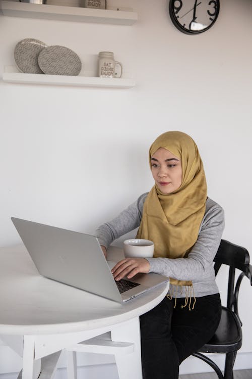 Young Muslim female remote worker in casual wear typing on netbook while sitting at plastic table with cup of coffee in apartment