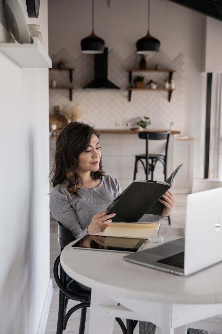 Ethnic Female Student Reading Copybook Near Laptop And Tablet