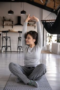 一位快乐的年轻少数族裔女性盘腿坐在公寓的瑜伽垫上，戴着耳机闭眼听音乐，并举起了手