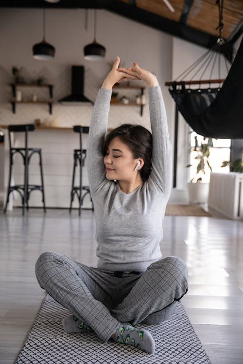 Cheerful young ethnic female in earbuds sitting on yoga mat with crossed legs and closed eyes while listening to music and raising hands in apartment