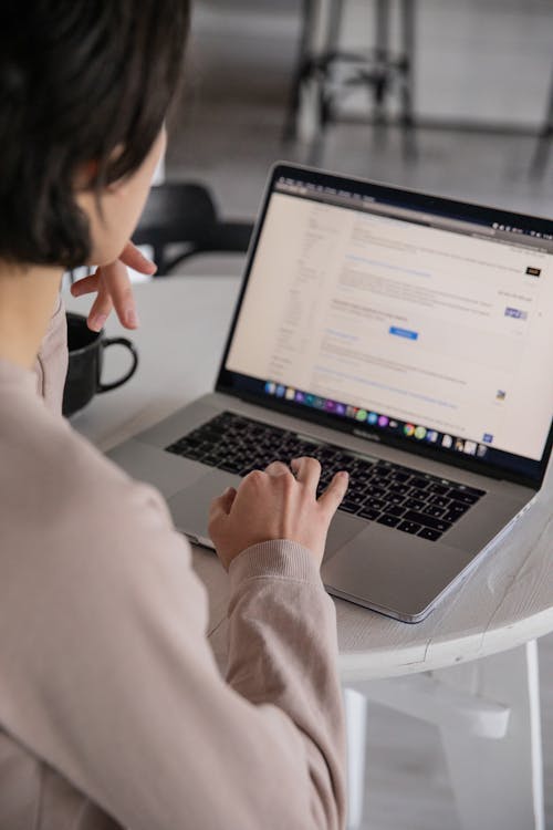 Free Back view of crop anonymous female remote employee typing on portable computer while watching website on screen and sitting at plastic table with cup of hot drink in apartment Stock Photo