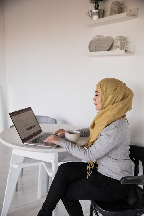 Side view of smiling young Muslim freelancer sitting with crossed legs and typing on portable computer while watching website on screen and drinking coffee in apartment