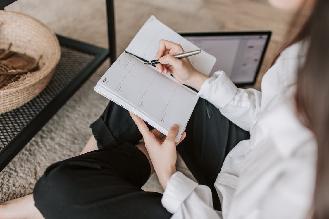 Side view of faceless woman in casual clothes taking notes on notepad while sitting in lotus pose on floor in modern apartment during daytime