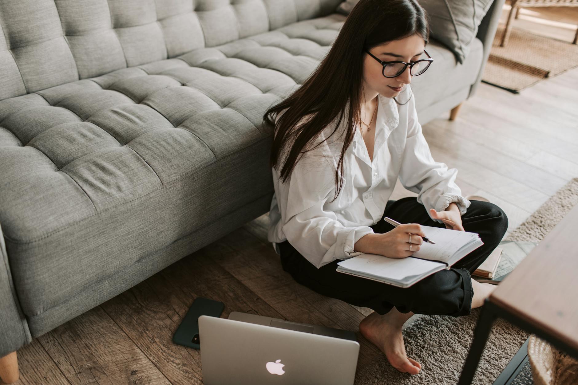 A young woman sitting on the floor using a laptop and writing notes in a cozy apartment.