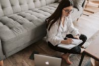 一名年轻女子坐在舒适的公寓地板上，用笔记本电脑写笔记