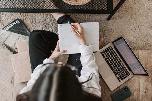 Top view of anonymous woman in casual wear sitting on floor with laptop and smartphone and creating plan on notebook while resting during break in modern living room
