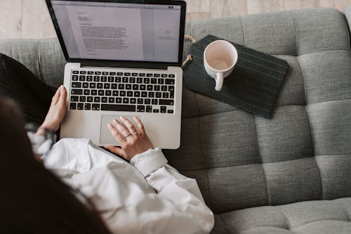 From above of woman in casual wear sitting on comfortable couch with cup and typing on keyboard of laptop while working remotely in cozy living room