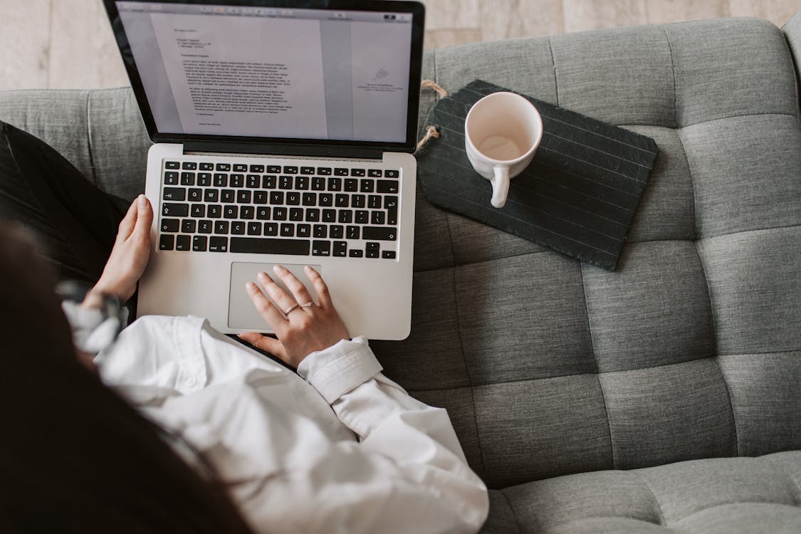 Free From above of woman in casual wear sitting on comfortable couch with cup and typing on keyboard of laptop while working remotely in cozy living room Stock Photo