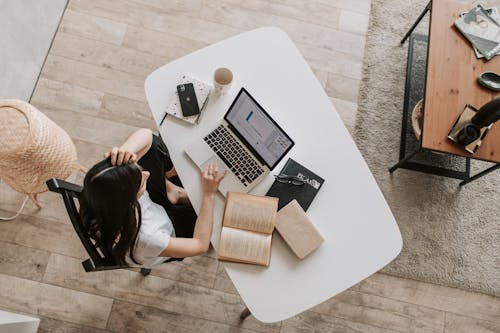 Free From above of young woman with long dark hair in casual clothes working at table and browsing netbook while sitting in modern workplace and touching hair Stock Photo