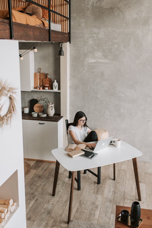 From above of serious young lady in casual wear typing on keyboard of laptop while sitting on chair at table with books and cup of coffee in comfortable living room