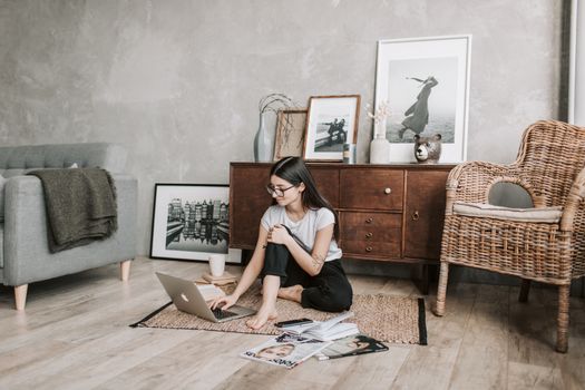 Positive female in casual clothes and eyeglasses reading netbook while sitting barefoot on floor carpet in modern living room with minimalist interior in eco style during free time
