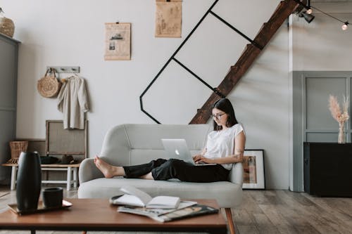Free Positive female with tattooed arms wearing casual clothes and eyeglasses reading netbook while sitting barefoot on comfortable sofa behind table with books and magazines in modern apartment with minimalist interior in eco style during free time Stock Photo