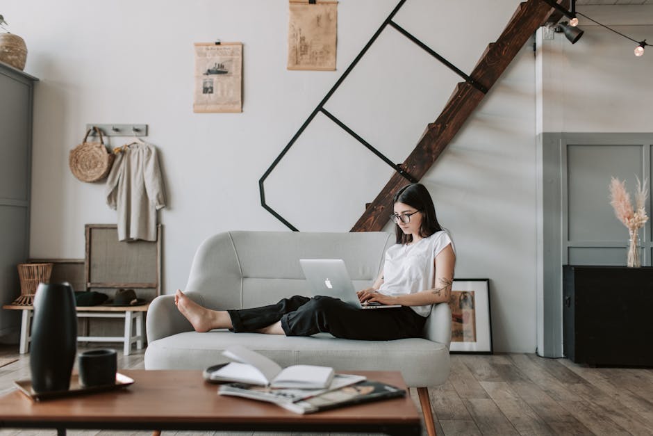 Positive female with tattooed arms wearing casual clothes and eyeglasses reading netbook while sitting barefoot on comfortable sofa behind table with books and magazines in modern apartment with minimalist interior in eco style during free time