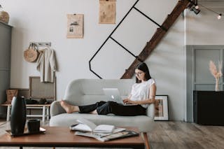 Positive female with tattooed arms wearing casual clothes and eyeglasses reading netbook while sitting barefoot on comfortable sofa behind table with books and magazines in modern apartment with minimalist interior in eco style during free time
