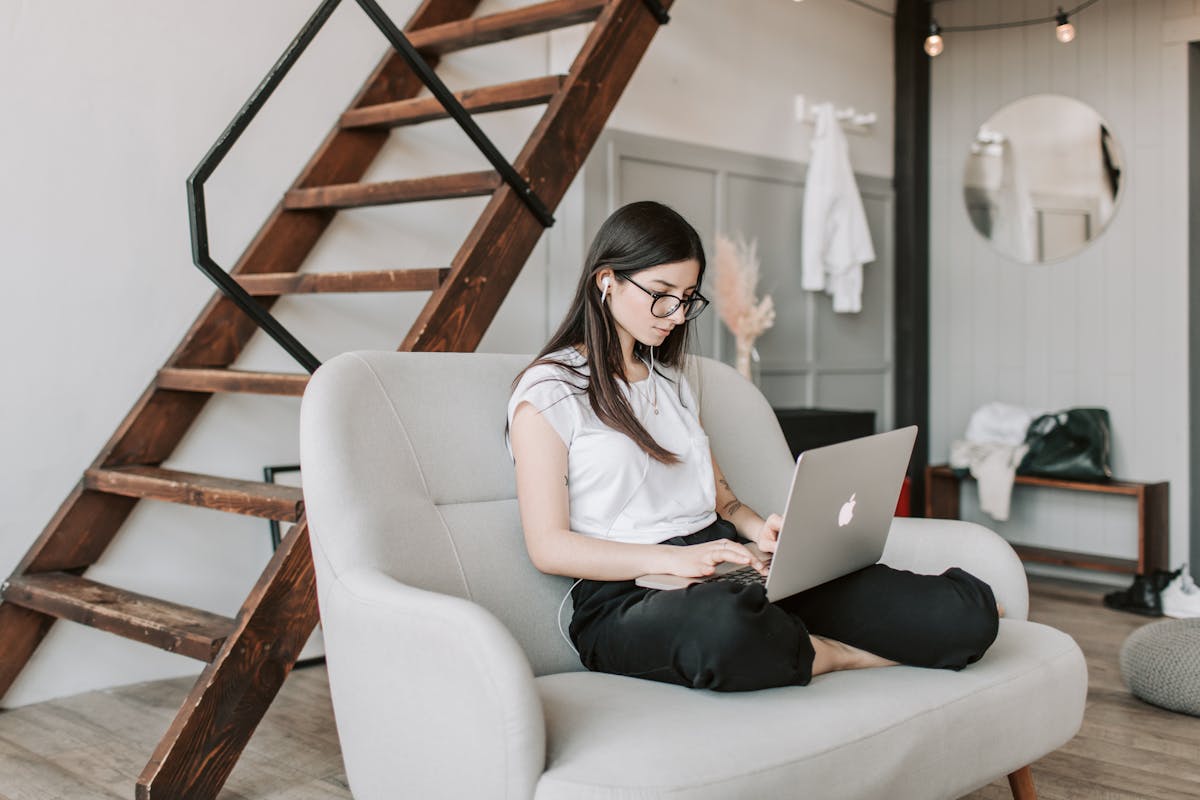 Young female in eyeglasses and casual clothes sitting barefoot on sofa in modern apartment with wooden staircase while wearing earbuds and watching movie on netbook