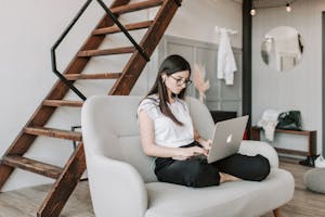Young female in eyeglasses and casual clothes sitting barefoot on sofa in modern apartment with wooden staircase while wearing earbuds and watching movie on netbook