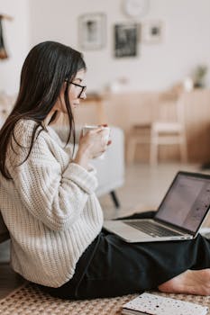 A young woman in a cozy setting works on her laptop while enjoying a cup of tea.
