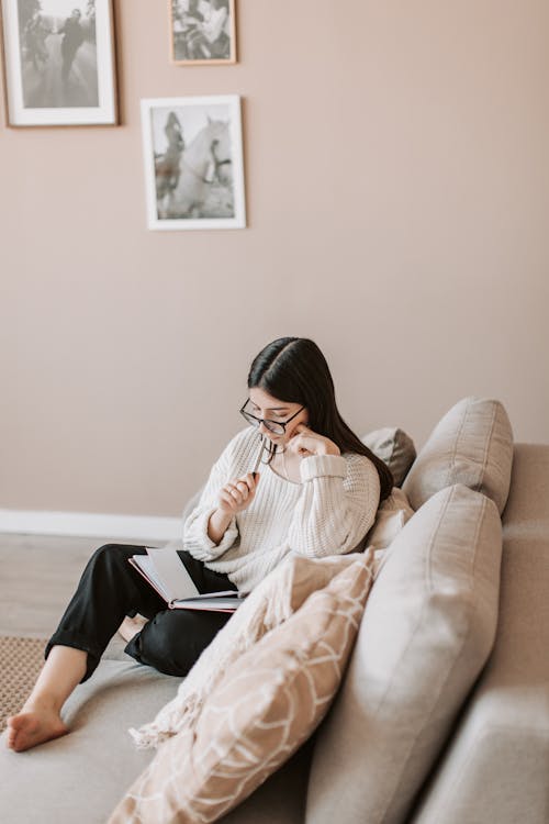 Free Concentrated businesswoman sitting barefoot with legs crossed on comfortable sofa in modern living room and planning business project during remote work while touching cheek and lips with pen Stock Photo