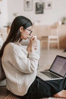 A woman in a cozy sweater works from home on a laptop while enjoying tea.
