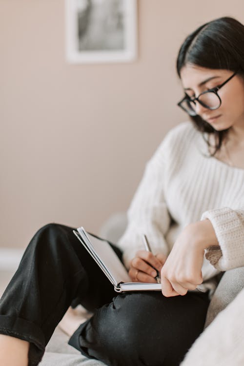 Free Attentive female wearing eyeglasses and casual outfit sitting barefoot with crossed legs on comfortable couch in modern flat and taking notes in notebook with pen Stock Photo