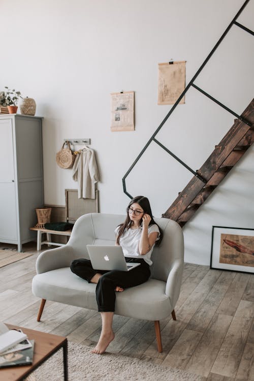 Full body barefoot female with tattooed arms wearing casual clothes and eyeglasses browsing netbook while sitting on comfortable sofa in modern apartment during weekend