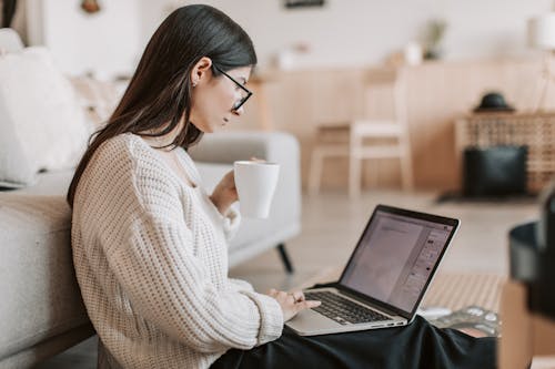 Free Side view of female freelancer in warm sweater and eyeglasses drinking tea from white ceramic cup while sitting on floor near sofa with netbook on legs while creating document for remote work project Stock Photo