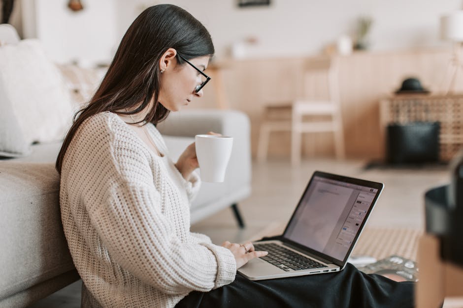 Side view of female freelancer in warm sweater and eyeglasses drinking tea from white ceramic cup while sitting on floor near sofa with netbook on legs while creating document for remote work project