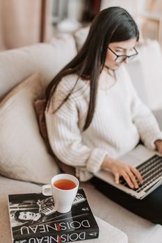 From above of focused female in casual wear sitting on sofa with cup of hot drink placed on book while spending time at home