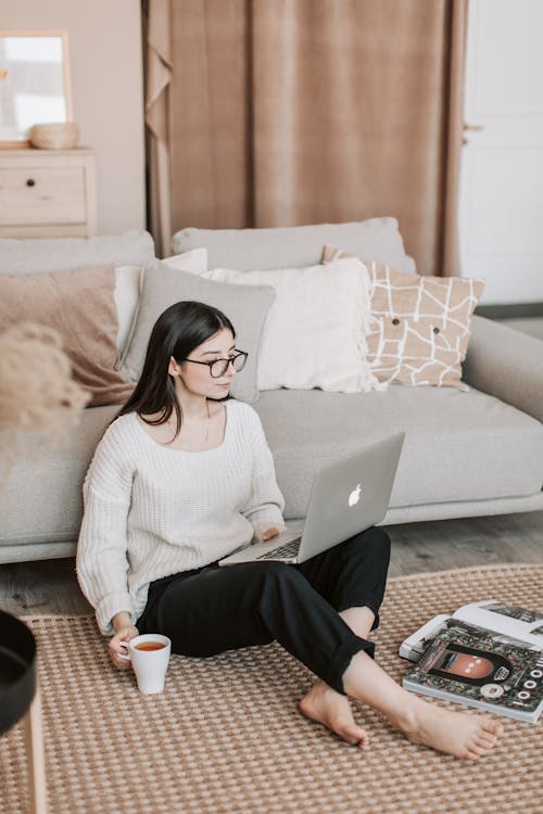 Free Focused female in casual wear sitting on floor carpet near sofa with cup of tea and browsing laptop while spending time at home Stock Photo