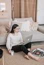 Focused female in casual wear sitting on floor carpet near sofa with cup of tea and browsing laptop while spending time at home