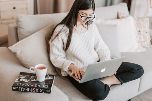 From above crop female in casual clothes and eyeglasses sitting on cozy sofa with book and cup of tea while using laptop for surfing Internet