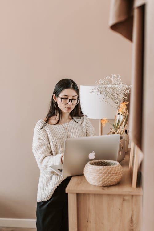 Free Concentrated young female in trendy casual outfit using laptop while standing near cabinet in cozy living room of modern apartment Stock Photo