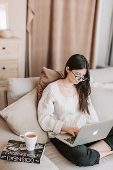 From above of young female in casual clothes and eyeglasses using laptop browsing Internet while sitting on comfortable sofa near book and cup of tea in modern apartment