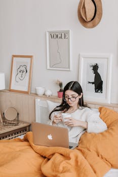 A woman relaxing in bed with coffee and laptop, surrounded by art and comfort.