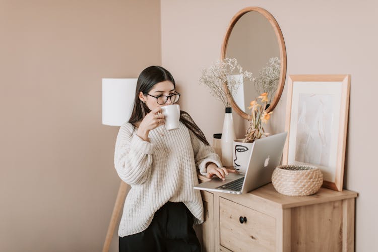 Busy Female Using Laptop And Drinking Hot Beverage At Home