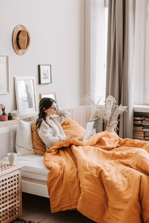 Free Side view of young female in warm sweater sitting on bed under blanket using laptop at cozy home apartment Stock Photo