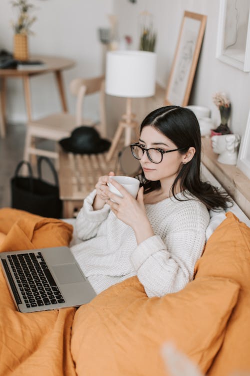 Dreamy brunette in glasses enjoying hot drink while cuddling under blanket on bed with laptop near